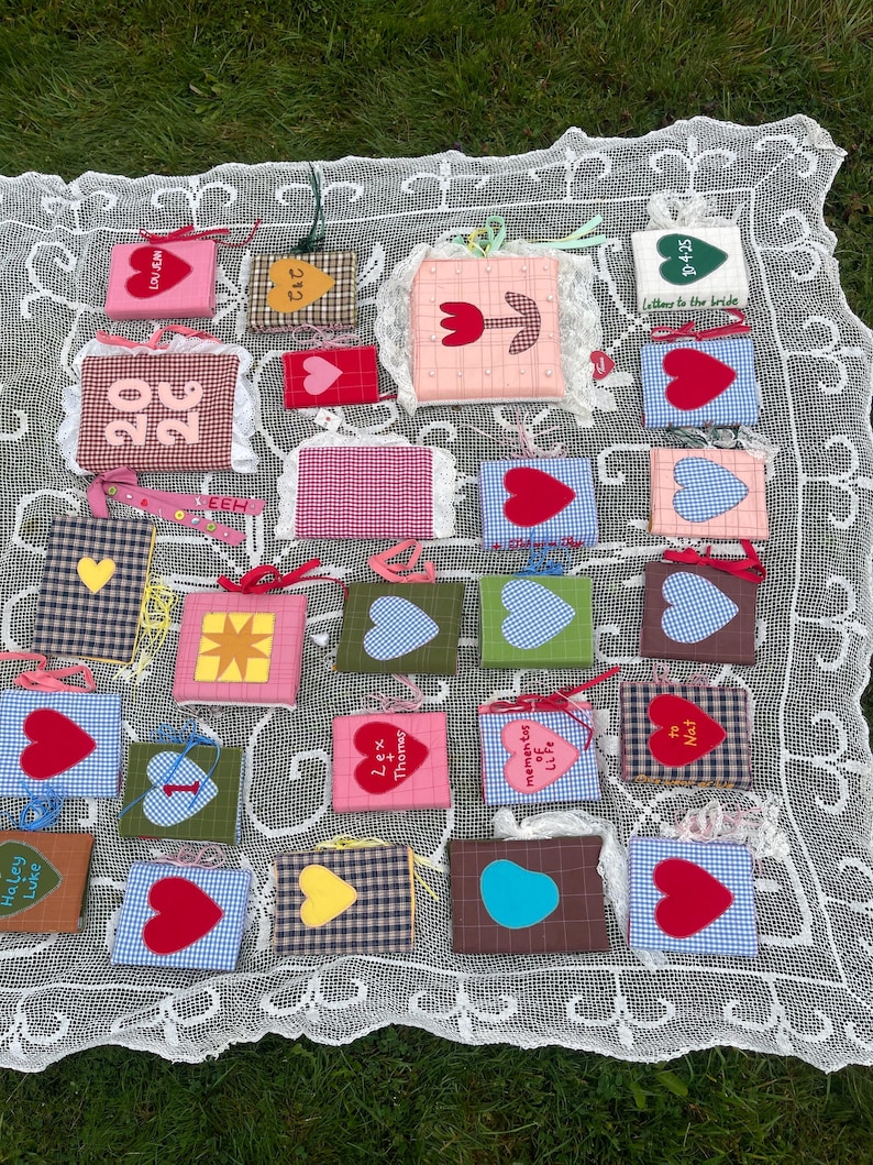Peut inclure: Une collection de petits objets carr&eacute;s en tissu avec divers motifs, notamment des c&oelig;urs, des &eacute;toiles et des motifs floraux. Certains objets comportent du texte comme "LOVE" et des noms. Les objets sont dispos&eacute;s sur une nappe en dentelle blanche.