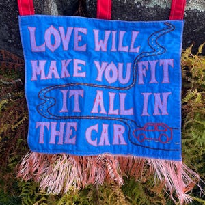 以下が含まれることがあります： 「LOVE WILL MAKE YOU FIT IT ALL IN THE CAR」の刺繍が施された青いバナー。 バナーは道路と車のデザインで、下部にピンクのフリンジ、上部に吊り下げ用の赤いタブが付いています。