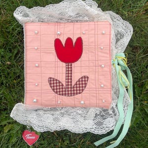 Könnte beinhalten: Ein rosa gesteppter Buchumschlag mit einem roten Tulpen-Applikation und Perlenakzenten. Der Umschlag ist mit weißer Spitze verziert und mit hellgrünen und gelben Bändern gebunden. Ein herzförmiger Anhänger mit dem Wort "Texas" hängt daran.