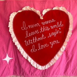 Könnte beinhalten: Ein pinkfarbener Wandbehang mit einem großen roten Herz in der Mitte. Das Herz trägt die weiße Aufschrift: „I never wanna leave this world without sayin' I love you“. Das Herz ist mit weißer Spitze verziert und der untere Teil hat rosa Fransen.