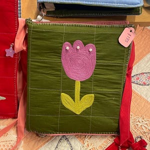 May include: A green quilted crossbody bag with a pink tulip applique and yellow stem and leaves. The bag has a pink lining and strap, and a tag that reads £175. Other bags are visible in the background.