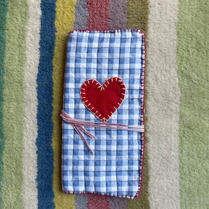 Könnte beinhalten: Ein handgefertigtes Notizbuch mit blau-weißem Vichy-Muster und einem roten Herz-Applikation. Das Notizbuch ist mit einem rosa Band gebunden und hat rote Nähte an den Rändern. Der Einband ist gesteppt.