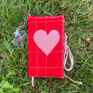 May include: A red fabric journal with a pink heart applique and a silver envelope charm. The journal has a grid pattern and a pink ribbon closure. The charm is attached with a silver chain. The journal is lying on green grass.