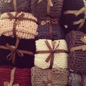 Puede incluir: Una pila de mantas de punto grueso de varios colores, cada una atada con una cinta marrón y una pequeña etiqueta. Las mantas están dispuestas en forma de pirámide, con la manta superior de color rosa.