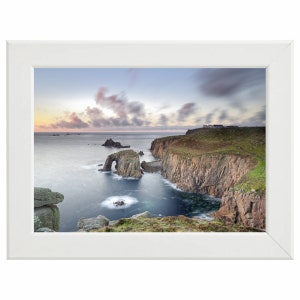 May include: A panoramic view of a rocky coastline with a natural arch formation. The ocean is a deep blue with white waves crashing against the rocks. The sky is a soft pink and purple with white clouds.