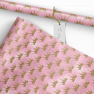 May include: Pink wrapping paper with a repeating pattern of gold glitter crowns. The paper has a subtle shimmer.