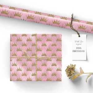 May include: Pink wrapping paper with a gold glitter crown pattern. The paper is wrapped around a gift box with a brown twine bow. A gift tag with the text "With Love on your 10th BIRTHDAY" is attached to the twine.