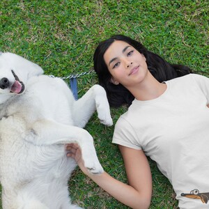 Pu&ograve; includere: Una donna con lunghi capelli neri giace sulla schiena su un prato verde con un husky bianco. La donna indossa una maglietta bianca con un bassotto di cartone animato stampato su di essa. Il cane &egrave; sdraiato sulla schiena con le zampe in aria.