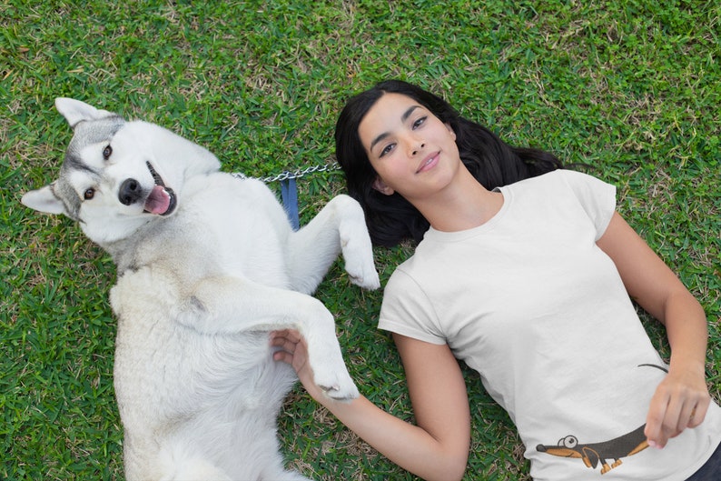 Pu&ograve; includere: Una donna con lunghi capelli neri giace sulla schiena su un prato verde con un husky bianco. La donna indossa una maglietta bianca con un bassotto di cartone animato stampato su di essa. Il cane &egrave; sdraiato sulla schiena con le zampe in aria.