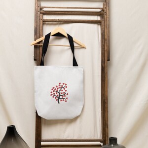 Puede incluir: Bolsa de lona blanca con asas negras colgada de un estante de madera. La bolsa presenta un dise&ntilde;o de &aacute;rbol de corazones rojos.