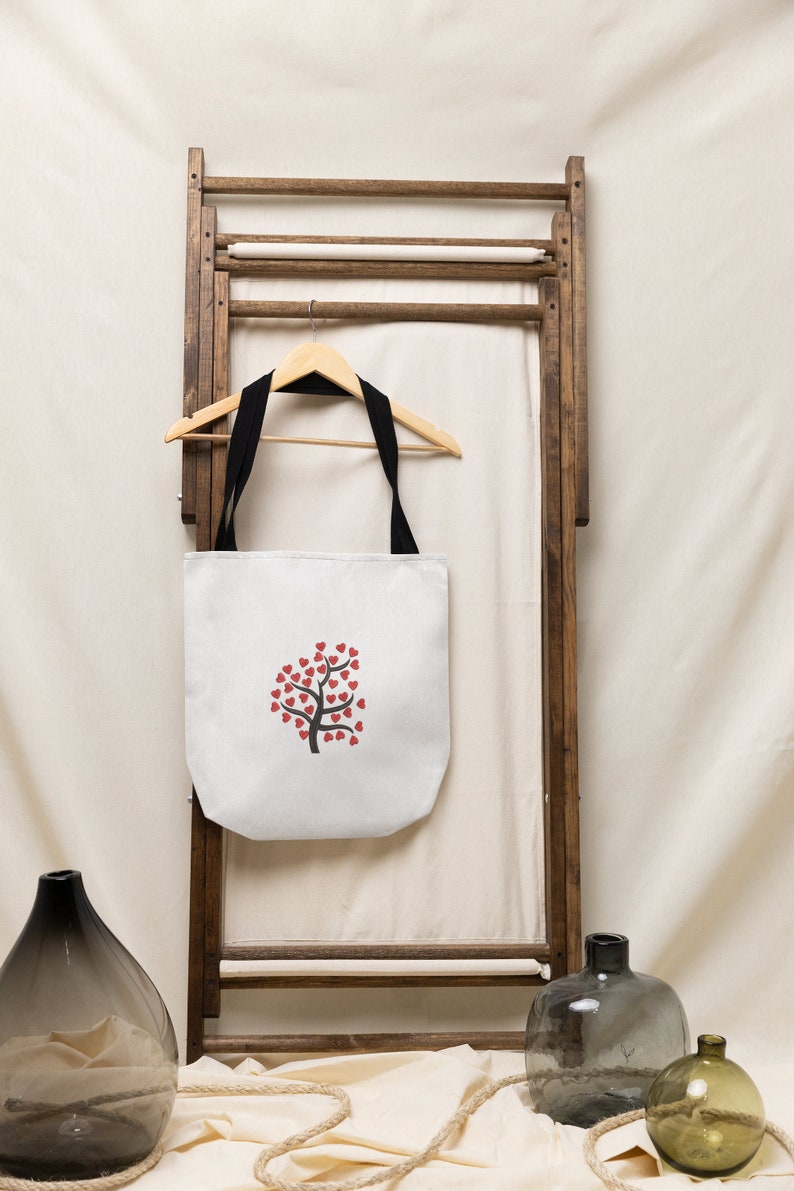 Puede incluir: Bolsa de lona blanca con asas negras colgada de un estante de madera. La bolsa presenta un dise&ntilde;o de &aacute;rbol de corazones rojos.
