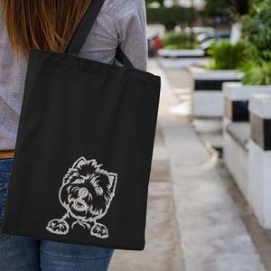 K&ouml;nnte beinhalten: Eine schwarze Tragetasche mit einem wei&szlig;en gestickten Bild eines Hundekopfes, der hinter der Tasche hervorlugt.