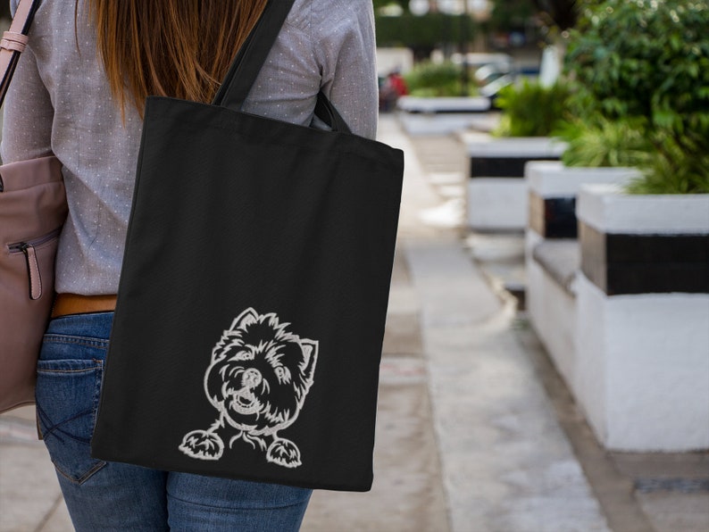 K&ouml;nnte beinhalten: Eine schwarze Tragetasche mit einem wei&szlig;en gestickten Bild eines Hundekopfes, der hinter der Tasche hervorlugt.