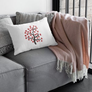 Puede incluir: Una almohada blanca con un dise&ntilde;o de &aacute;rbol de corazones rojos en un sof&aacute; gris con una manta de color rosa.