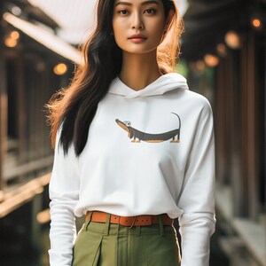 Pu&ograve; includere: Una donna indossa una felpa con cappuccio bianca con un disegno di un bassotto dei cartoni animati. Indossa una cintura marrone e pantaloni verdi.