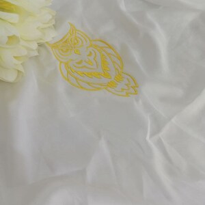 Puede incluir: Tela blanca con un dise&ntilde;o de b&uacute;ho bordado en amarillo.