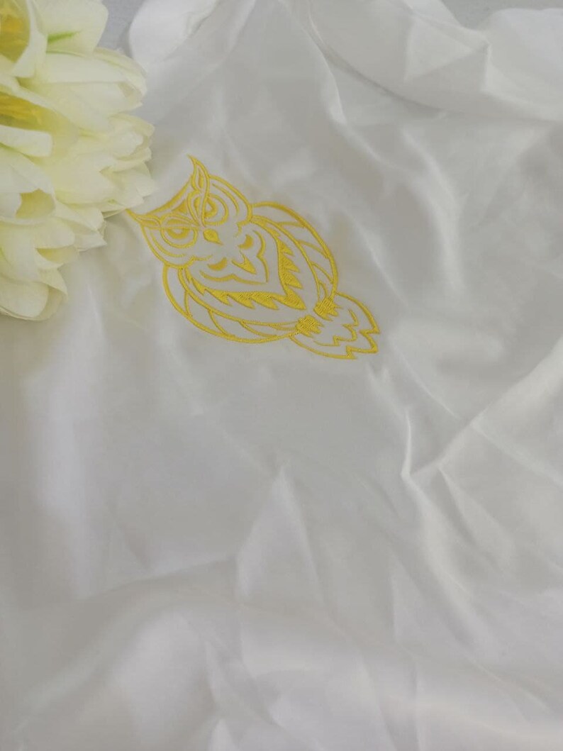 Puede incluir: Tela blanca con un dise&ntilde;o de b&uacute;ho bordado en amarillo.