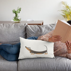Pu&ograve; includere: Un cuscino bianco con un'illustrazione di un cartone animato di un bassotto marrone e nero con occhi grandi. Il bassotto &egrave; disteso con la testa inclinata verso l'alto.