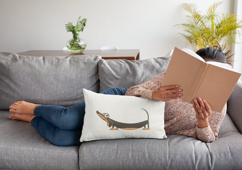 Pu&ograve; includere: Un cuscino bianco con un'illustrazione di un cartone animato di un bassotto marrone e nero con occhi grandi. Il bassotto &egrave; disteso con la testa inclinata verso l'alto.