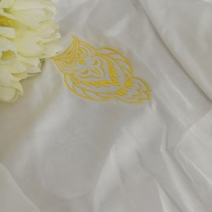 Puede incluir: Tela blanca con un dise&ntilde;o de b&uacute;ho bordado en amarillo.