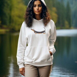 Pu&ograve; includere: Una felpa con cappuccio bianca con un bassotto di cartone animato stampato sul davanti. La modella indossa la felpa e si trova in piedi davanti a un lago.