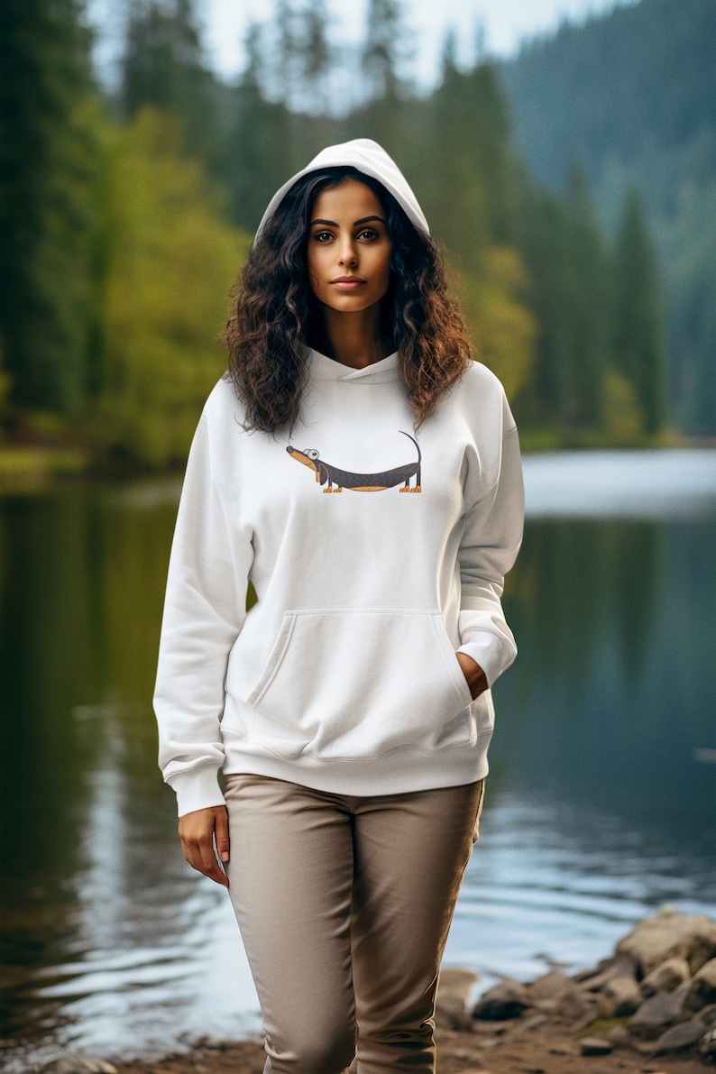 Pu&ograve; includere: Una felpa con cappuccio bianca con un bassotto di cartone animato stampato sul davanti. La modella indossa la felpa e si trova in piedi davanti a un lago.