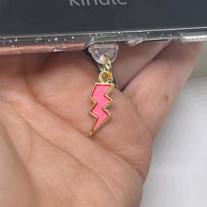 Puede incluir: Un dije de rayo rosa con borde dorado y un pequeño anillo dorado. El dije está sujeto a una funda transparente para teléfono.