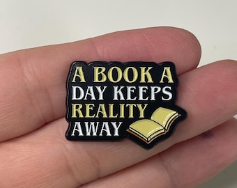 May include: Black enamel pin with the text "A BOOK A DAY KEEPS REALITY AWAY" in white and gold lettering. The pin features an open book illustration in yellow and black. A literary-themed accessory.