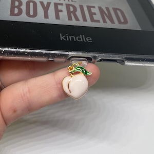 Könnte beinhalten: Ein goldener Pfirsich-Anhänger mit grünen Blättern, der an einem Kindle E-Reader befestigt ist. Der Kindle zeigt den Text "The Boyfriend" und "kindle" an.