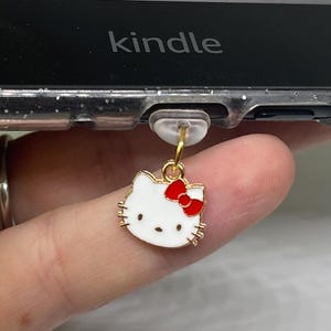May include: A gold charm with a white Hello Kitty design and a red bow attached to a clear phone case with the word "kindle" printed on it.