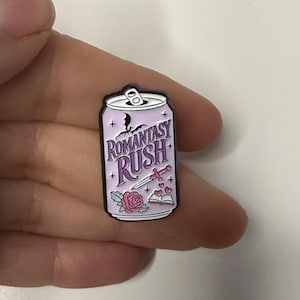 May include: A lavender enamel pin in the shape of a soda can. The pin features the words "Romantasy Rush" in a stylized font, along with illustrations of a rose, a dagger, an open book, and a bat silhouette.