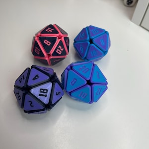May include: Four multi-colored, geometric dice with numbers. The dice are in shades of purple, blue, and pink. Each die has a different color scheme and number arrangement, suitable for tabletop games.