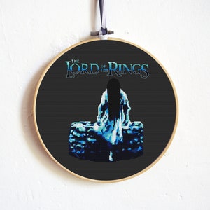 May include: A black and white cross-stitch embroidery hoop with a blue and white image of a woman in a white dress sitting on a stone wall. The text "The Lord of the Rings" is above the image.