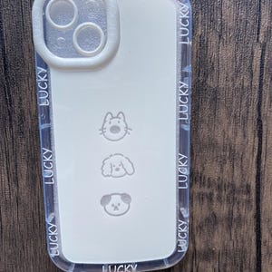 Peut inclure: Coque de téléphone transparente avec un intérieur blanc orné de trois visages d'animaux de dessin animé et le mot "LUCKY" répété sur les bords. La zone de l'objectif de l'appareil photo est blanche.