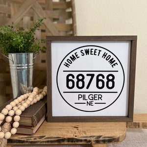 May include: A white wooden sign with a black circle and the text "Home Sweet Home" and the number "68768" with the text "Pilger NE" below the number.