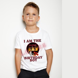 May include: White t-shirt with the text "I AM THE BIRTHDAY BOY" in red and black. The shirt features a graphic of a ninja character from the Ninjago series. The shirt is short-sleeved and has a crew neck.
