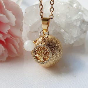 Könnte beinhalten: Eine goldfarbene Halskette mit einem strukturierten, kugelförmigen Anhänger. Der Anhänger hat ein Lebensbaum-Design mit kleinen, klaren Steinen. Ein kleiner weißer Stein und eine goldene Kette vervollständigen die Halskette.