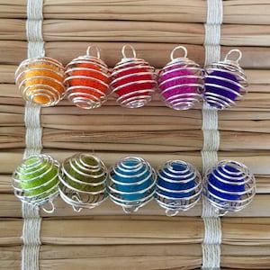 Pode incluir: Um conjunto de dez bolas coloridas cobertas de feltro, encerradas em gaiolas de arame prateado. As bolas estão dispostas em arco-íris, cada bola de uma cor diferente.