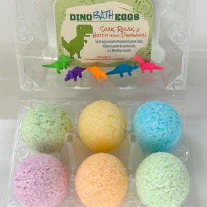 Può includere: Un contenitore di plastica trasparente con sei bombe da bagno colorate in tonalità pastello di verde, giallo, blu, viola, arancione e verde. Il contenitore contiene anche piccoli giocattoli di dinosauri di plastica e una carta con "Dino Bath Eggs".