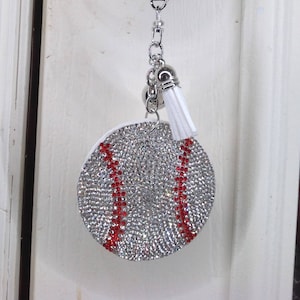 May include: A baseball-themed keychain hanging from a gold door knob. The baseball charm is covered in silver rhinestones with red stitching. A white tassel hangs from the bottom of the charm.