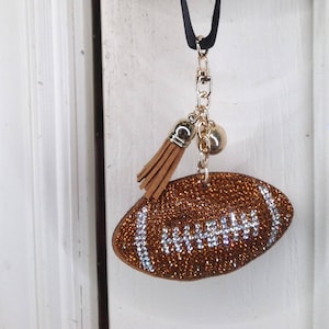 May include: A brown and white football-shaped door hanger with rhinestones. The hanger has a gold chain and a brown tassel.