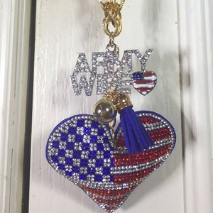 Może przedstawiać: Złoty łańcuszek z sercowatym wisiorkiem, na którym napis "Army Wife" wykonany jest z cyrkonii. Serce ozdobione jest czerwonymi, białymi i niebieskimi cyrkoniami w wzorze amerykańskiej flagi. Wisiorek ma niebieski frędzl.