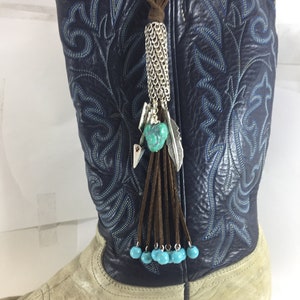 May include: A decorative boot tassel featuring a silver chain, turquoise beads, and a feather charm. The tassel is attached to a brown suede cord and hangs from a navy blue and cream-colored cowboy boot. The boot has intricate blue stitching.