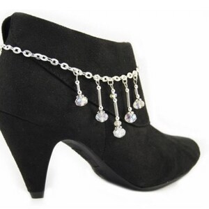 May include: Black ankle boot with a silver chain and dangling crystal accents. The boot has a pointed heel and a suede-like texture. The chain is draped around the ankle, with multiple crystal charms hanging down.
