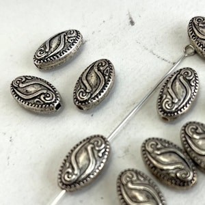 May include: A group of silver oval-shaped beads with a decorative design. The beads have a raised, ornate pattern and a smooth, polished finish.