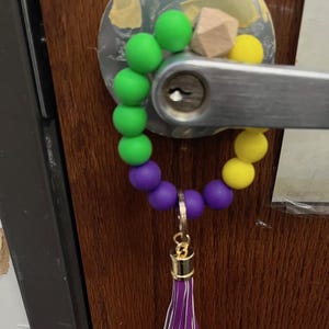 May include: A beaded keychain wristlet in green, purple, and yellow, with a gold-colored clasp and a purple tassel. The keychain is attached to a door handle.