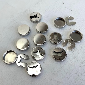 Peut inclure: Un ensemble de boutons-pression argentés, assemblés et démontés, disposés sur une surface claire. Les attaches comprennent des capuchons ronds et plats et des anneaux de retenue fendus séparés. Ils sont probablement utilisés pour des vêtements ou des projets d'artisanat.