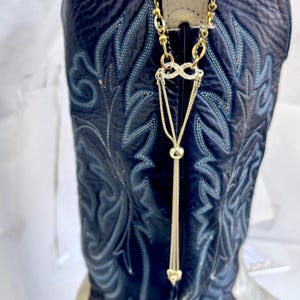 Könnte beinhalten: Nahaufnahme eines marineblauen Cowboy-Stiefels mit aufwendigen hellblauen Nähten. Eine goldfarbene Kette mit Unendlichkeitszeichen und Quaste hängt am Zugriemen des Stiefels und verleiht dem Western-Stil einen Hauch von Eleganz.