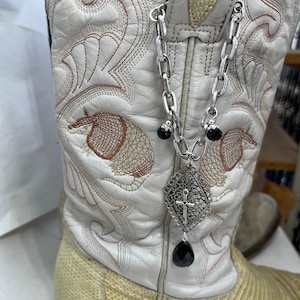 May include: White cowboy boot with intricate embroidery and a silver chain boot jewelry. The jewelry features a diamond-shaped pendant with a cross and a black teardrop-shaped bead. The boot has a snakeskin-like texture on the lower part.