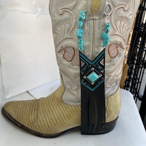 Può includere: Uno stivale da cowboy color crema con cuciture intricate e un collo del piede strutturato marrone chiaro. Un accessorio per stivali in pelle turchese e nera con frange pende dallo stivale, aggiungendo un tocco decorativo.